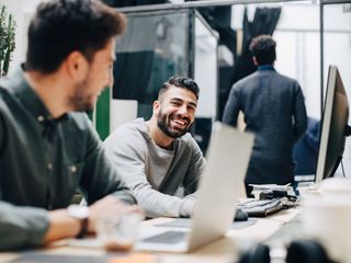 Smiling male colleagues looking at each other while sitting by desk in office
