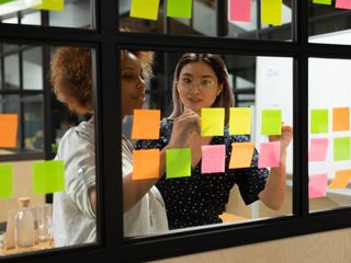 Two women putting notes on sticky paper at window glass agile kanban board at office