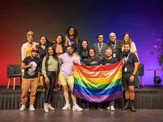 Foto de 14 pessoas, algumas delas seguram uma bandeira com as cores do arco &iacute;ris. 