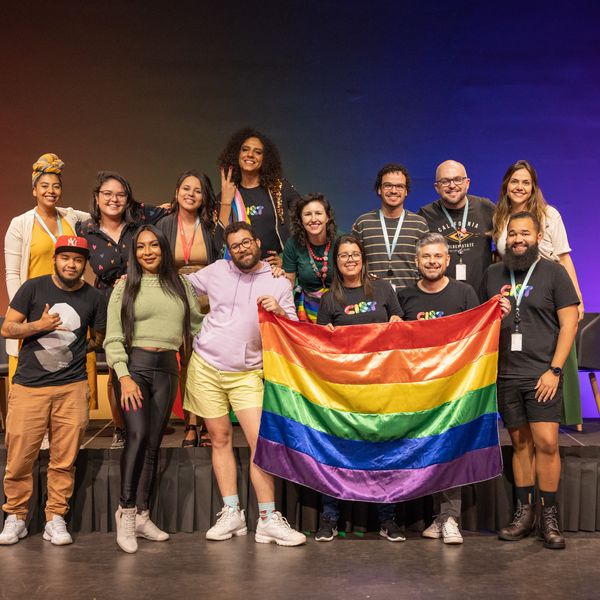 Foto de 14 pessoas, algumas delas seguram uma bandeira com as cores do arco &iacute;ris. 