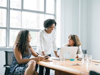 Foto de tr&ecirc;s mulheres conversando em um ambiente fechado. Duas est&atilde;o sentadas e a frente est&aacute; um notebook. 