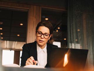Foto de mulher branca, sentada, em frente ao computador, observando algo em seu caderno &agrave; esquerda