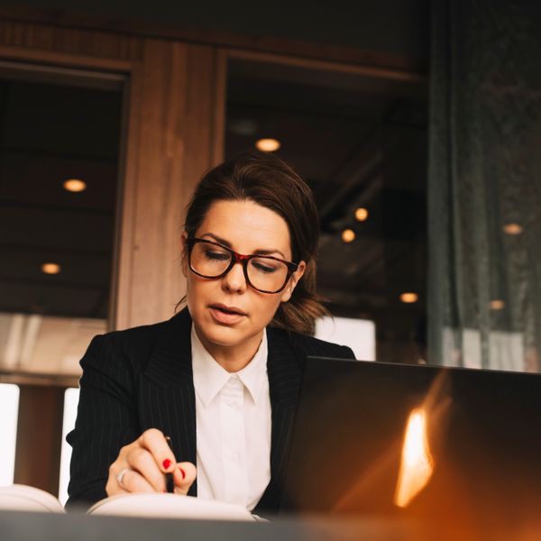 Foto de mulher branca, sentada, em frente ao computador, observando algo em seu caderno &agrave; esquerda
