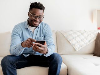 Black man at home texting on his smartphone while sitting on the sofa.