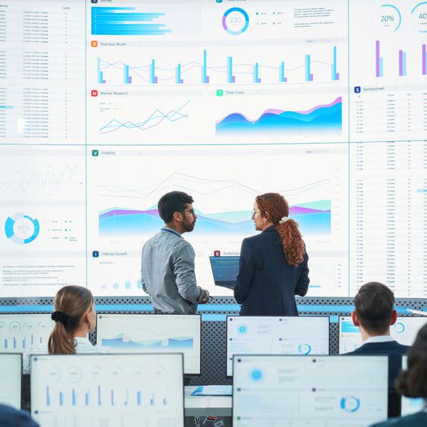 Back View Of Indian Data Analyst And Hispanic Manager Discussing Reports On Digital Screen in Monitoring Room.