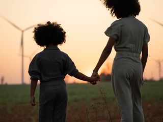 Crian&ccedil;a segura m&atilde;o de mulher ao fundo turbinas e&oacute;licas e por do sol