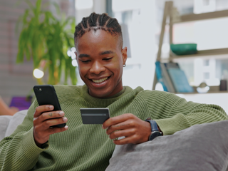 A young man sitting on a couch, using a smartphone in one hand and a credit card in the other.