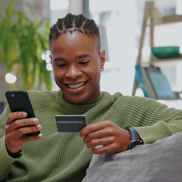 A young man sitting on a couch, using a smartphone in one hand and a credit card in the other.
