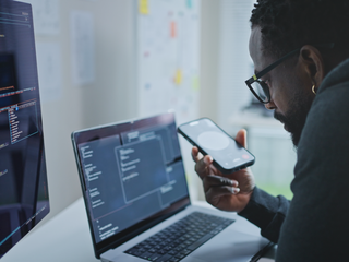 A person sitting at a desk using AI via smartphone while looking at a laptop and a monitor displaying coding.