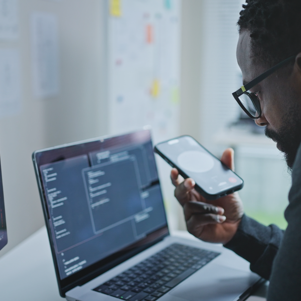 A person sitting at a desk using AI via smartphone while looking at a laptop and a monitor displaying coding.