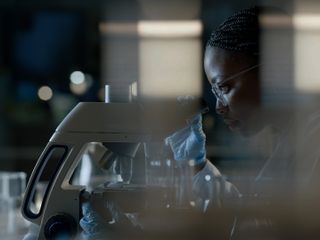 A person in a lab coat and blue gloves is examining a sample under a microscope in a dimly lit laboratory setting.