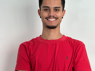 Lucas, homem pardo, de cabelo e barba pretos, sorrindo. Ele veste uma camisa vermelha.