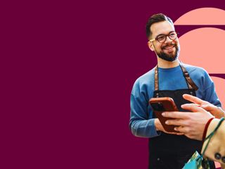 A bearded man with glasses wearing a black apron smiles as he assists a customer holding a phone. The burgundy background features a geometric design with peach circles.