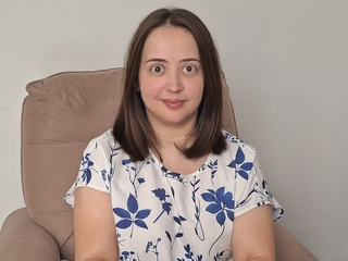 Jaqueline, mulher branca de cabelo castanho liso e olhos castanhos, est&aacute; sorrindo e veste uma camiseta branca com estampa de flores azuis.