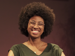 Gabriela, mulher negra de cabelo cacheado e olho preto, usando &oacute;culos e sorrindo. Ela veste uma blusa verde escuro.