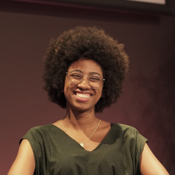Gabriela, mulher negra de cabelo cacheado e olho preto, usando &oacute;culos e sorrindo. Ela veste uma blusa verde escuro.