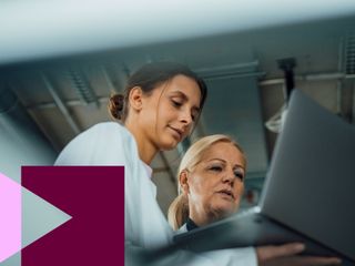 two women in lab coats reviewing information on a laptop in industrial setting