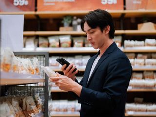 A person in a dark blazer is holding a phone and examining a snack package while shopping in a grocery store aisle filled with various packaged snacks on the shelves.