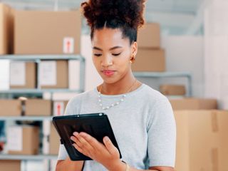 A woman standing in a warehouse using a tablet to manage inventory, with cardboard boxes and metal shelving in the background.