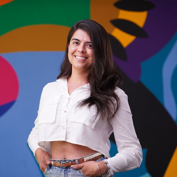 Julia, mulher branca de cabelo liso preto at&eacute; a altura dos ombros, veste blusa de manga comprida branca e sorri. Ao fundo, uma parede colorida.
