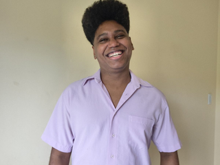 Antonio Lucas, homem negro de cabelo cacheado preto e olhos pretos, sorrindo. Ele veste uma camisa de bot&atilde;o lil&aacute;s.