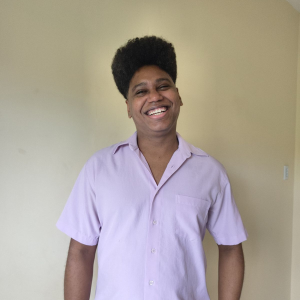 Antonio Lucas, homem negro de cabelo cacheado preto e olhos pretos, sorrindo. Ele veste uma camisa de bot&atilde;o lil&aacute;s.