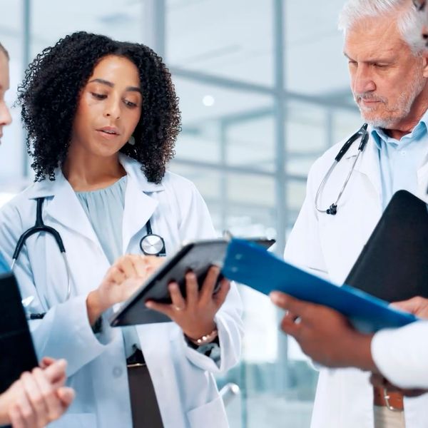 A diverse team of healthcare professionals in scrubs and white coats review patient information on a digital tablet in a bright hospital hallway.