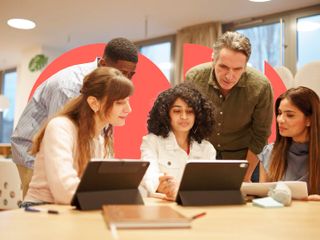 A diverse group of five colleagues collaborating around a table with tablets in a modern office, framed by large coral-colored geometric shapes in the background.