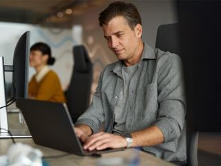 A man in a grey shirt focuses on his laptop at a modern office desk, with a colleague and other workstations visible in the blurred background.