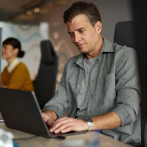A man in a grey shirt focuses on his laptop at a modern office desk, with a colleague and other workstations visible in the blurred background.