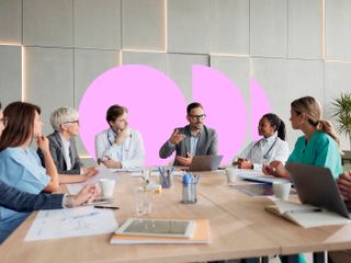 A man in a suit stands and presents to a diverse group of business and healthcare professionals seated around a large conference table.