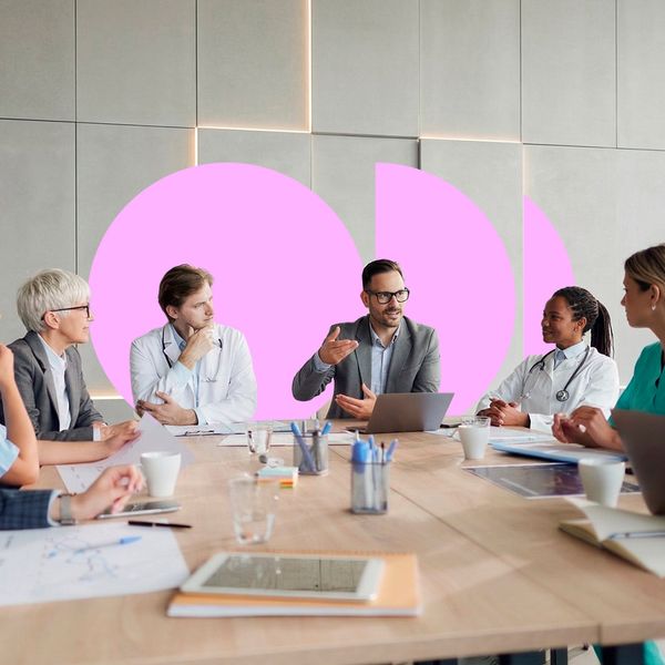 A man in a suit stands and presents to a diverse group of business and healthcare professionals seated around a large conference table.