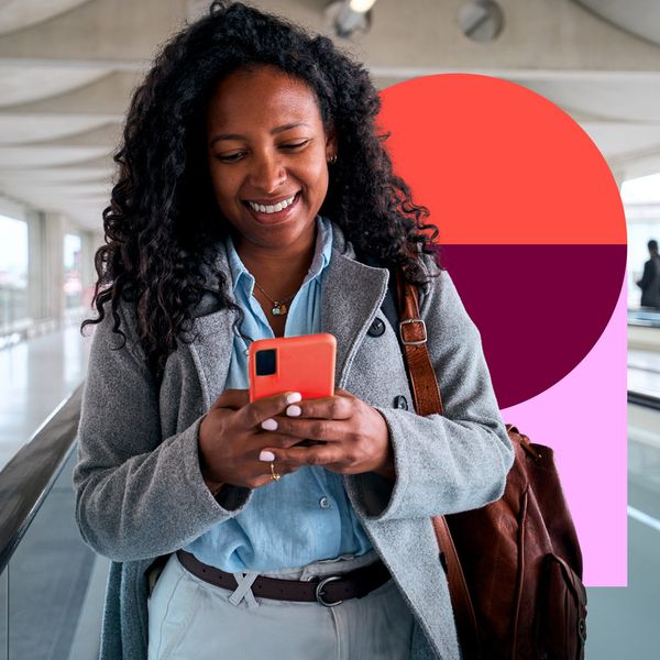 A smiling woman with curly hair looks at her smartphone while walking through a modern, bright transit terminal.