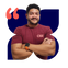 Acacio, homem pardo de cabelo cacheado preto, barba e bigode pretos, veste uma camisa bord&ocirc; e est&aacute; de bra&ccedil;os cruzados, sorrindo.