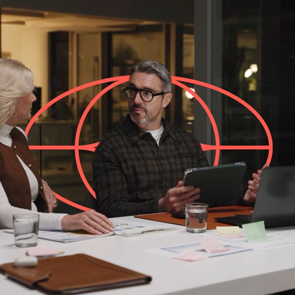 Three professionals engage in a focused meeting at a table with laptops and tablets in a modern office at night, featuring a red circular geometric overlay.