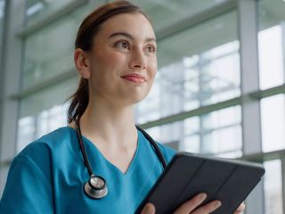 A smiling medical professional in blue scrubs holding a tablet in a bright, modern hospital setting.