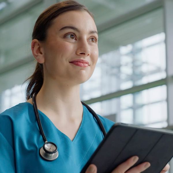 A smiling medical professional in blue scrubs holding a tablet in a bright, modern hospital setting.