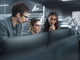 Three focused professionals lean in together to examine a computer monitor in a modern, high-tech workspace.