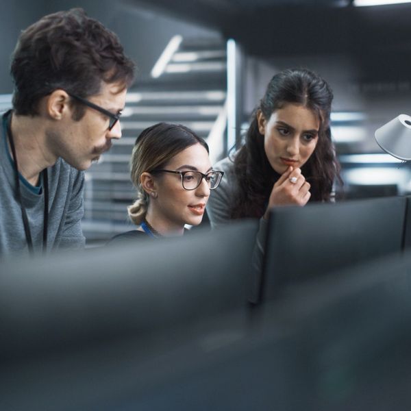 Three focused professionals lean in together to examine a computer monitor in a modern, high-tech workspace.