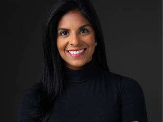 Ingrid, mulher parda de cabelo preto liso na altura dos ombros e olhos pretos, sorri. Ela est&aacute; de bra&ccedil;os cruzados e veste blusa preta.