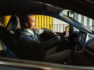 Side profile of a Black businessman in a suit driving a car equipped with a modern, glowing digital dashboard and infotainment screen.