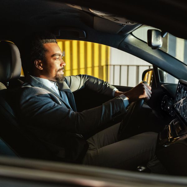 Side profile of a Black businessman in a suit driving a car equipped with a modern, glowing digital dashboard and infotainment screen.