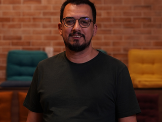 Domingos, homem branco de cabelo e barba pretos, usa &oacute;culos e camisa pretos. Est&aacute; sorrindo.