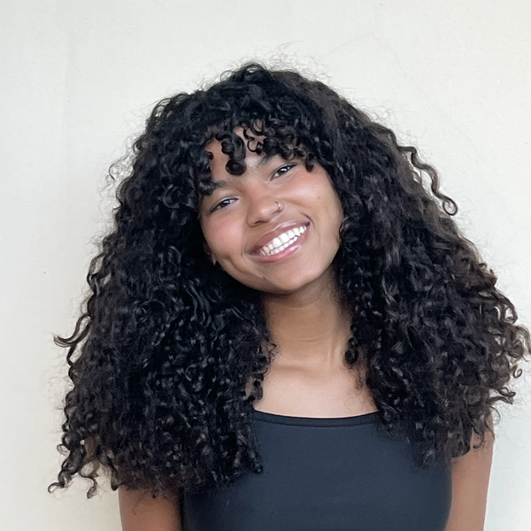 Eduarda, mulher parda de olhos pretos e cabelo cacheado preto na altura dos ombros com franja, em fundo azul-marinho. Ela sorri e veste uma blusa preta.