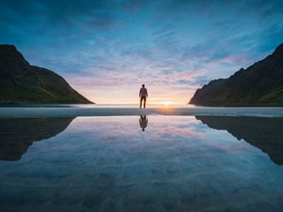 Person standing between mountains at sunset with reflections on the water