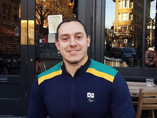 Daniel, homem branco de cabelo preto curto, sorrindo, veste um casaco azul marinho com a bandeira do Brasil e listras verde e amarelo.